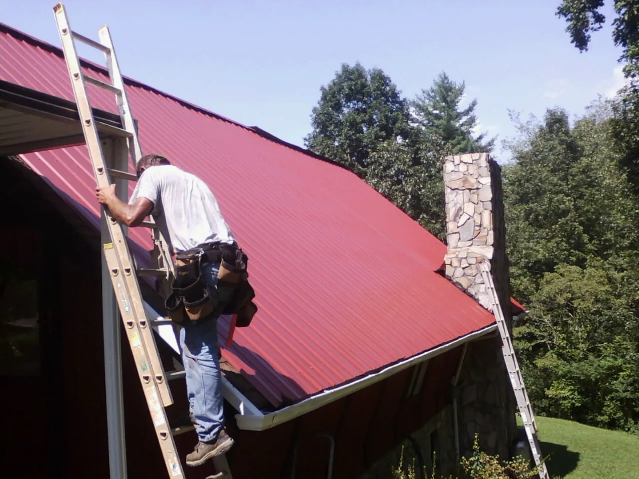 Roofer on a ladder beside a red metal roof and stone chimney — Soffit & Fascia Repair by Roofing Brothers in Gainesboro